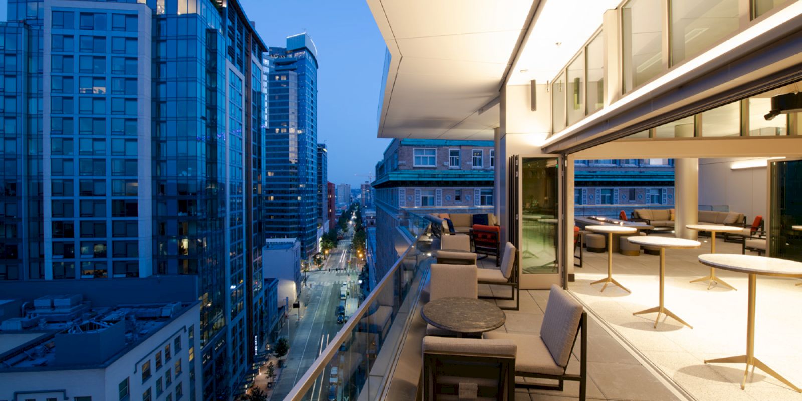 The image shows a modern balcony with chairs and tables overlooking a cityscape, featuring tall, illuminated buildings at dusk.