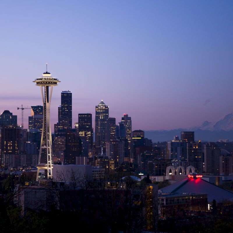 This is a twilight view of Seattle’s skyline, showcasing the Space Needle and Mount Rainier in the background, with city lights starting to twinkle.