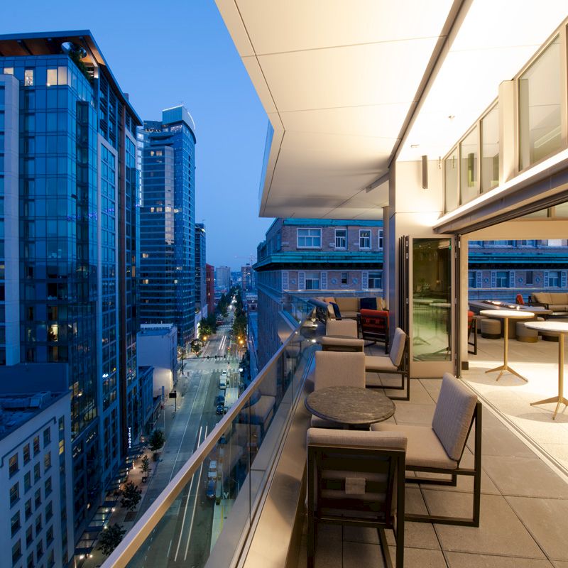 A modern cityscape at dusk viewed from a spacious balcony with seating and tables, adjacent to an illuminated indoor area.