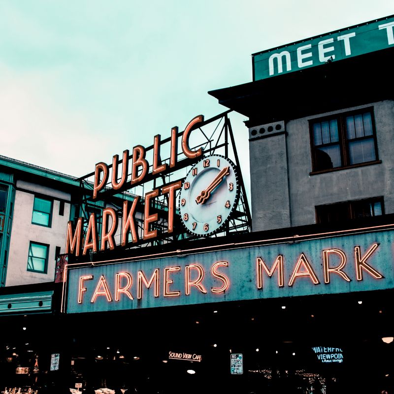 The image shows the iconic neon signs of a "Public Market" and "Farmers Market" with a clock, part of a well-known marketplace scene in an urban setting.