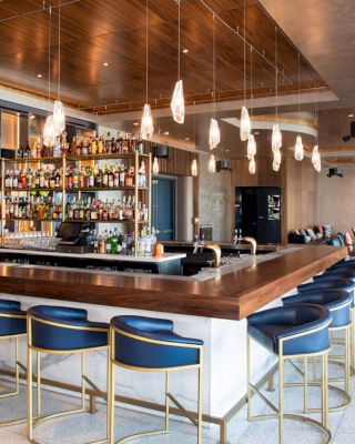 A modern bar with blue bar stools, high shelves with various bottles, hanging pendant lights, and a seating area with couches and chairs in the background.