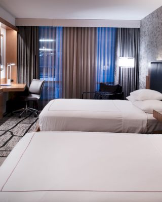 The image shows a modern hotel room with two neatly made beds, a wall-mounted TV, a workspace with a chair, and contemporary decor.
