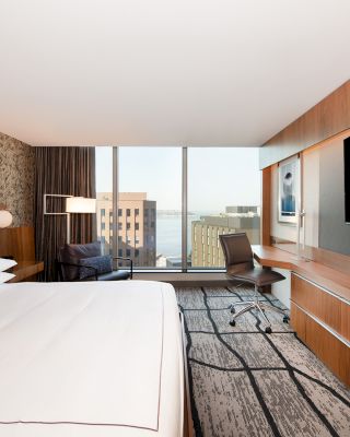 A modern hotel room features a large bed, a desk with a chair, a flat-screen TV, and a view of city buildings through a large window.