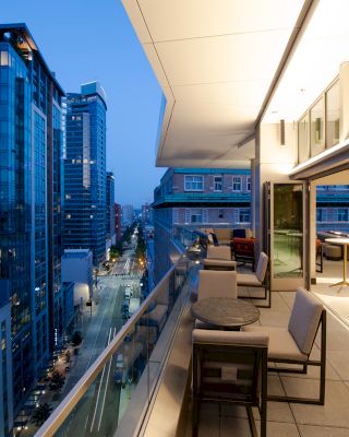 A modern balcony with seating and tables, overlooking an urban street surrounded by high-rise buildings during dusk.