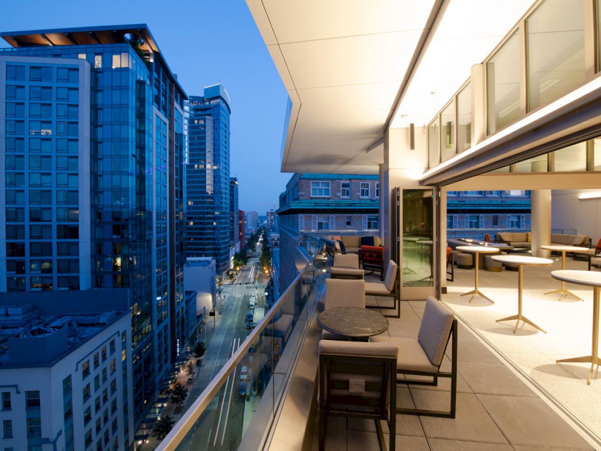 A modern urban balcony at dusk overlooking the city, with tables, chairs, and illuminated spaces inside and out.