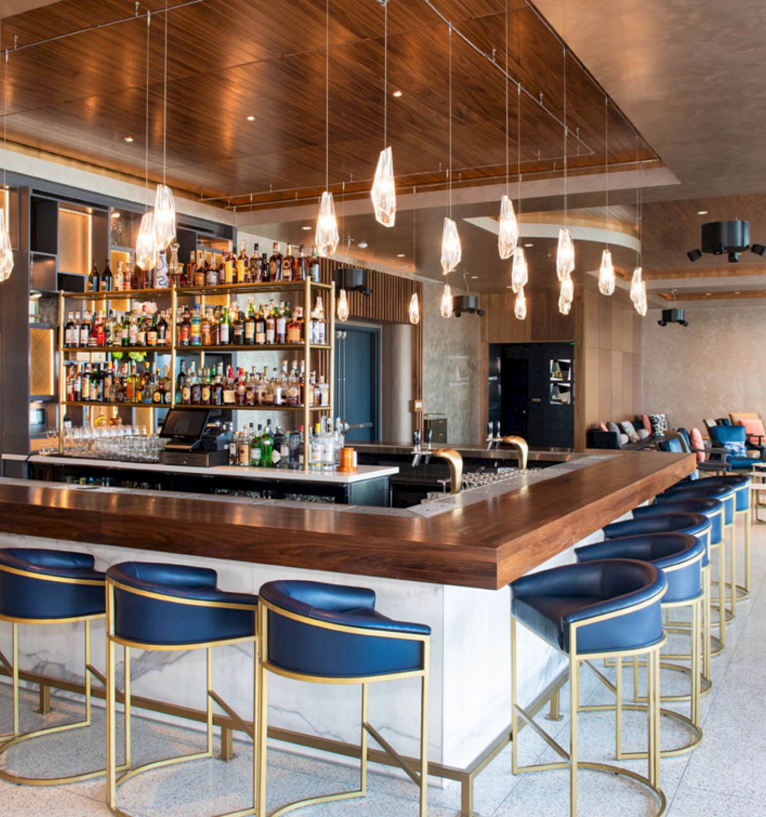 The image shows a modern bar with a wooden counter, blue bar stools, a well-stocked shelf of bottles, and hanging lights, in a contemporary setting.