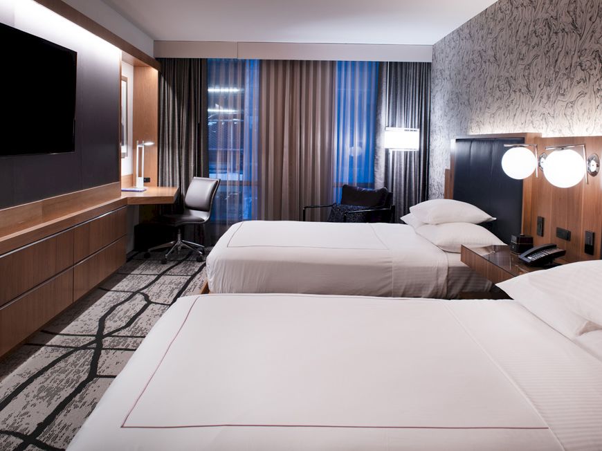 The image shows a modern hotel room with two double beds, a flat-screen TV, a desk, and contemporary decor, including stylish lamps and wallpaper.