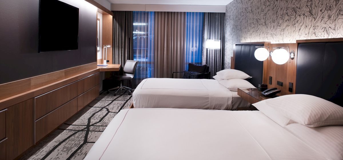 The image shows a modern hotel room with two double beds, a flat-screen TV, a desk, and contemporary decor, including stylish lamps and wallpaper.