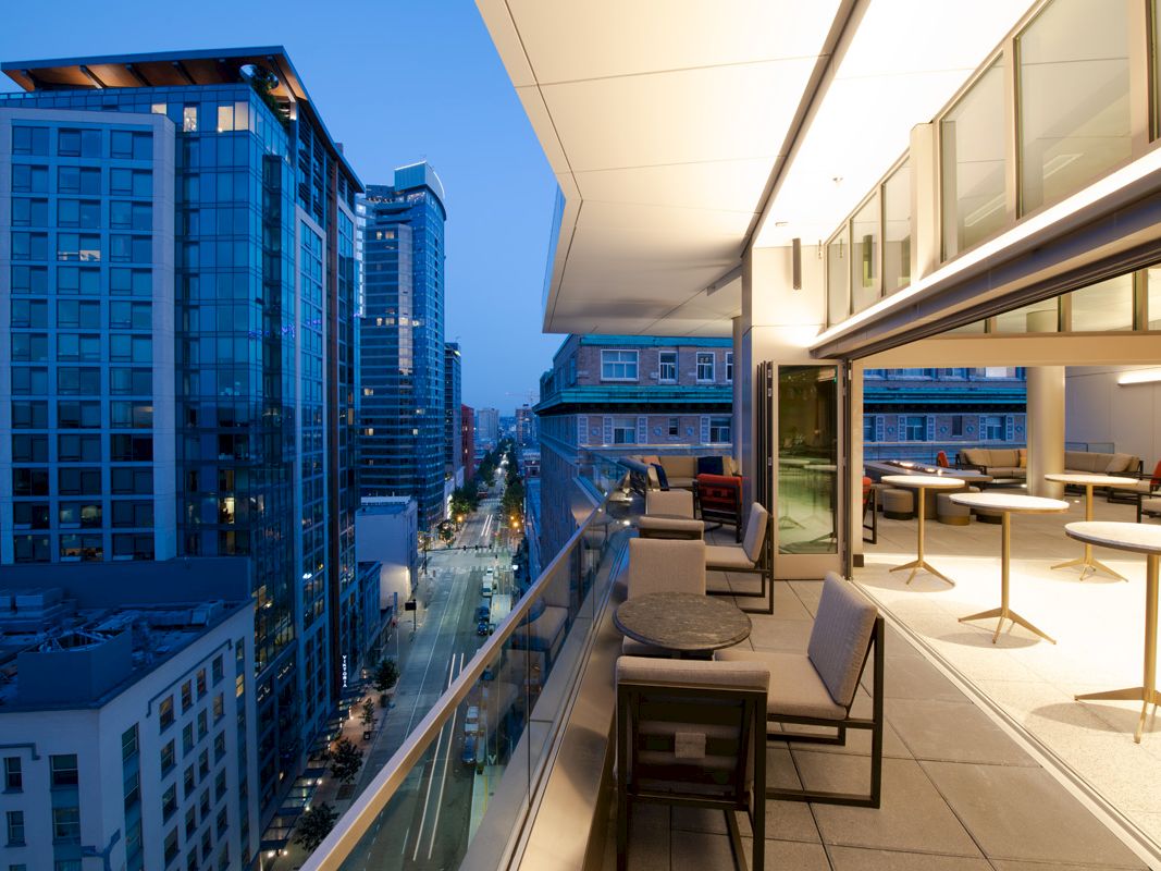 A modern balcony with chairs and tables overlooks a city street lined with tall buildings during dusk, with lights illuminating the area.