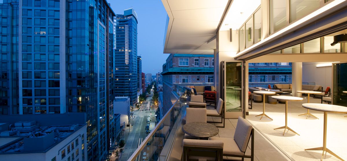 A modern balcony with chairs and tables overlooks a city street lined with tall buildings during dusk, with lights illuminating the area.