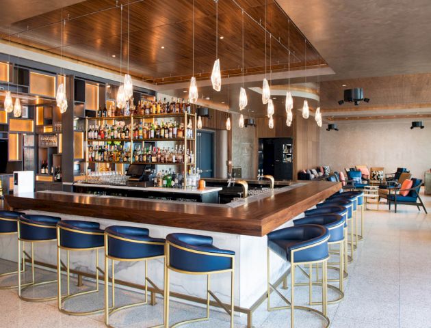 A modern bar with blue stools, a central wooden counter, hanging lights, and a well-stocked shelf of bottles, located in a spacious lounge area.