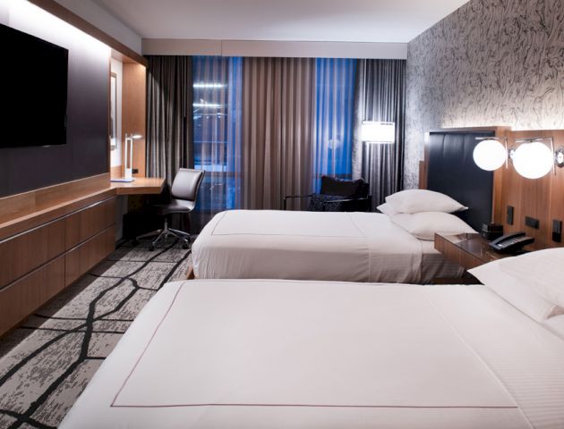 This image depicts a modern hotel room with two neatly-made beds, a wall-mounted TV, a desk, chair, and large windows with curtains.