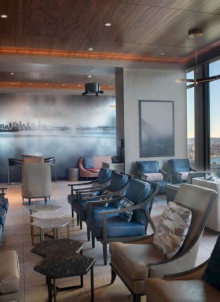 A modern lounge area with plush seating, a city skyline mural, large windows with waterfront views, and a Ferris wheel visible outside at the right.