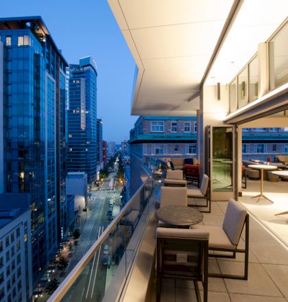 The image shows a modern balcony overlooking a cityscape at dusk. There are tables and chairs on the balcony with buildings visible in the background.