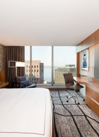 A modern hotel room with a large bed, desk, chair, wall-mounted TV, large window with a city view, and contemporary decor.