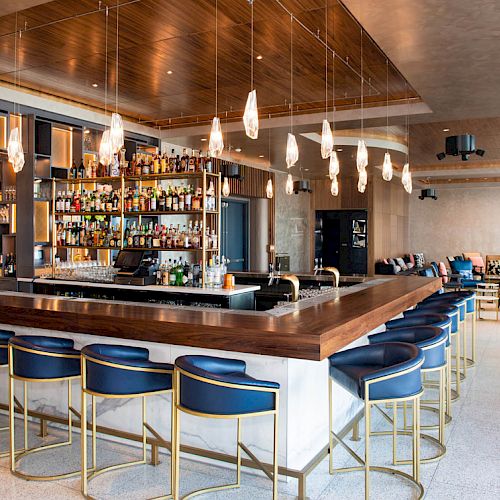 The image shows a modern bar with a wooden counter, blue chairs, and pendant lights, surrounded by bottles and glasses. The seating area is in the background.