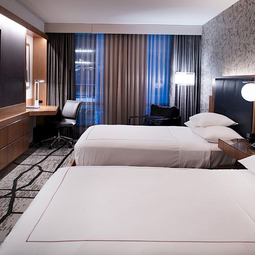 A modern hotel room with two beds, a TV, a desk, and chairs, featuring stylish lighting and wallpaper, creating a cozy and contemporary ambiance.