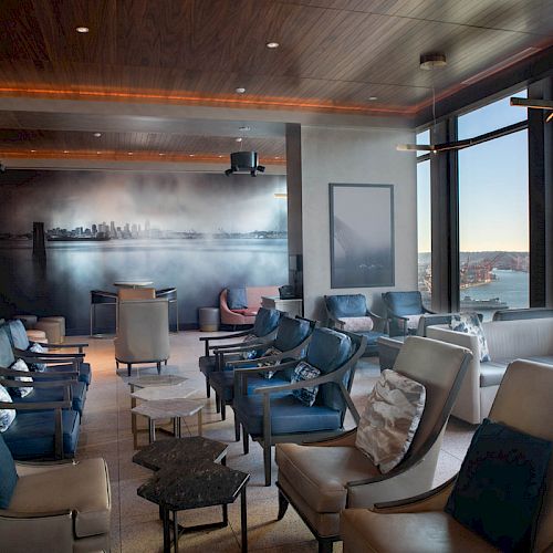 A modern lounge with numerous comfortable chairs, a mural on the wall, and large windows offering a scenic view of a waterfront and Ferris wheel.