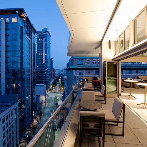 A modern balcony overlooks a busy city street lined with tall buildings. The balcony has seating and tables, with warm lighting inside.