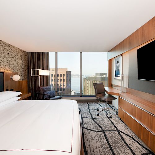 A modern hotel room with a large bed, flat-screen TV, desk and chair, and a window offering a city and water view, complemented by stylish decor.