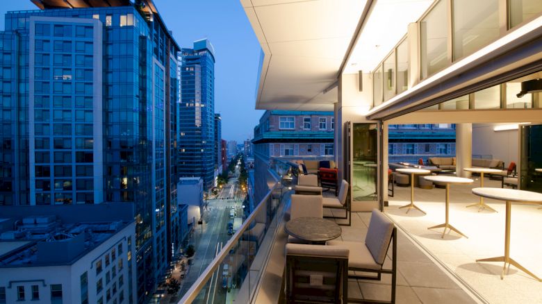 A modern urban balcony with seating, overlooking a city street and tall buildings at dusk. The view shows a mix of residential and commercial buildings.