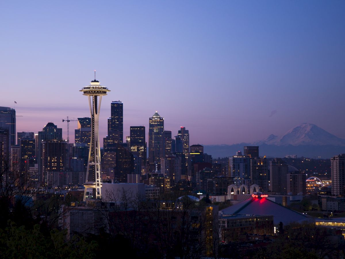 The image shows a nighttime city skyline with a prominent tower and distant mountains. Buildings are illuminated, and the sky has a purple hue at sunset.