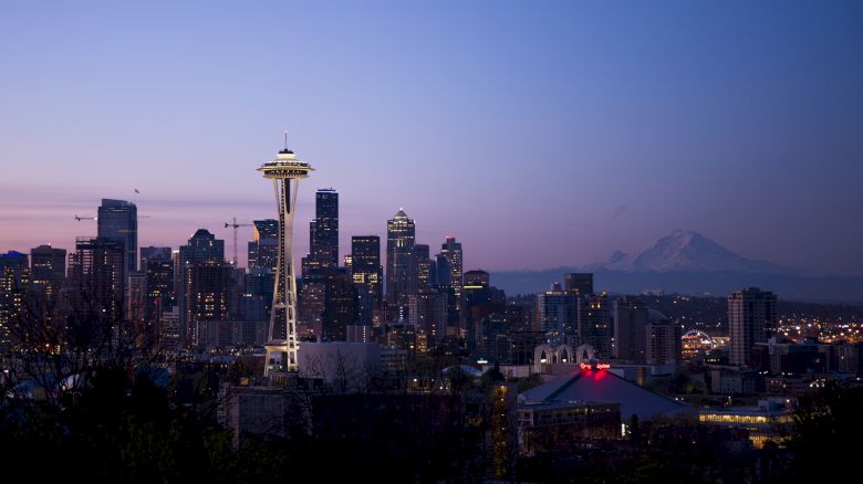 The image shows a nighttime city skyline with a prominent tower and distant mountains. Buildings are illuminated, and the sky has a purple hue at sunset.