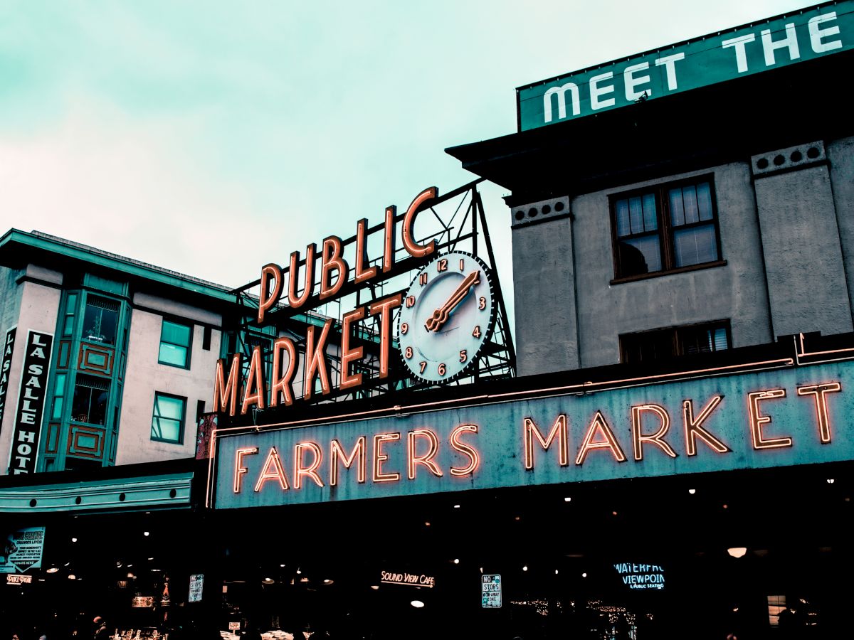 Public market with a neon "Farmers Market" sign, a clock, and surrounding buildings. It appears to be a popular market area bustling with activity.