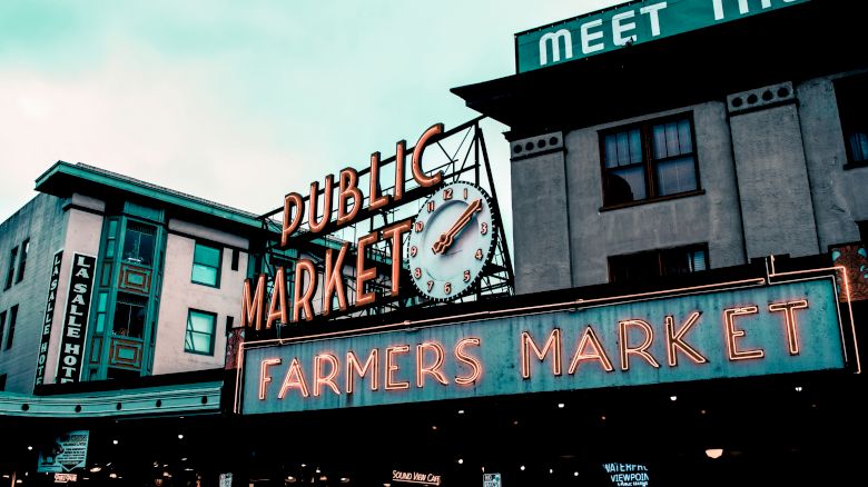 Public market with a neon "Farmers Market" sign, a clock, and surrounding buildings. It appears to be a popular market area bustling with activity.