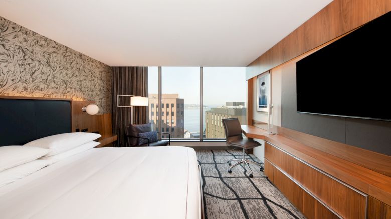 The image shows a modern hotel room with a large bed, a wall-mounted TV, a desk, and a chair. The window offers a view of buildings outside.