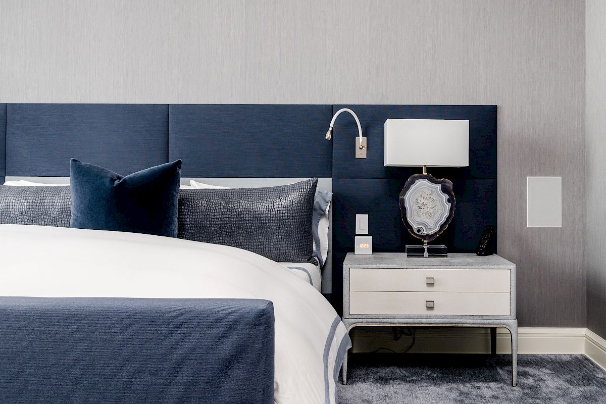 The image shows a modern bedroom with a blue upholstered bed, white and gray bedding, a nightstand with a lamp, and a decorative item.