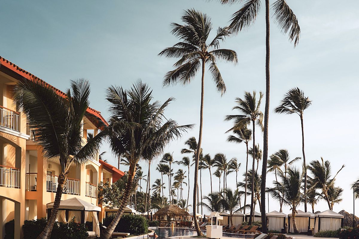 A serene poolside scene with lounge chairs, tall palm trees, and a building in the background under a clear blue sky.