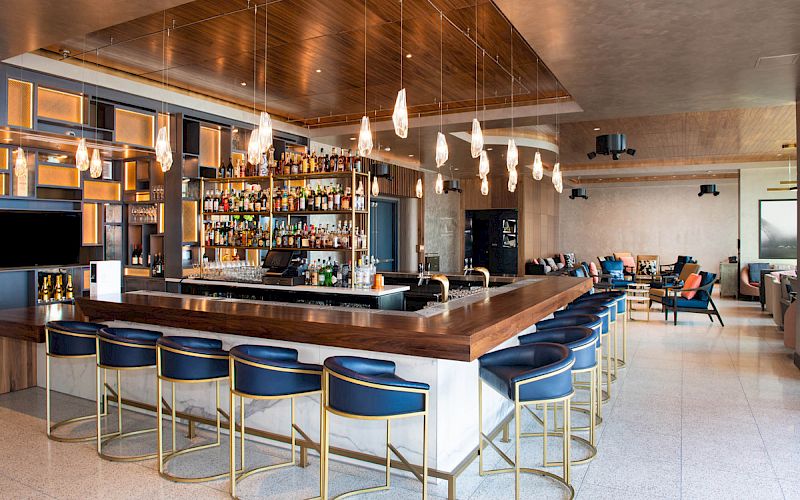 A modern bar with a wood counter, blue and gold stools, a shelf full of bottles, hanging lights, and a seating area in the background.
