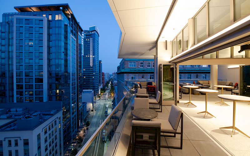 The image shows a modern balcony with tables and chairs overlooking a cityscape with tall buildings at dusk or evening.