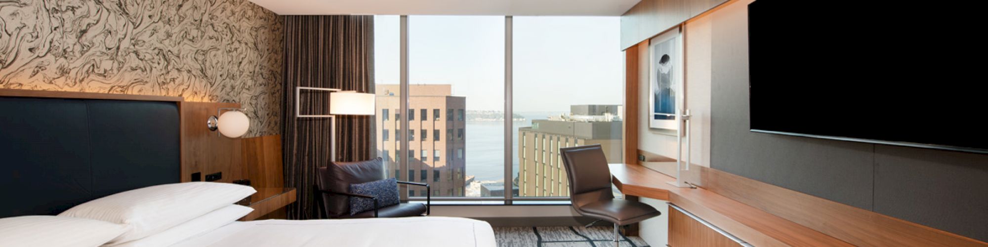 Modern hotel room with a large bed, wall-mounted TV, desk, and chair. Features floor-to-ceiling windows with a city view.
