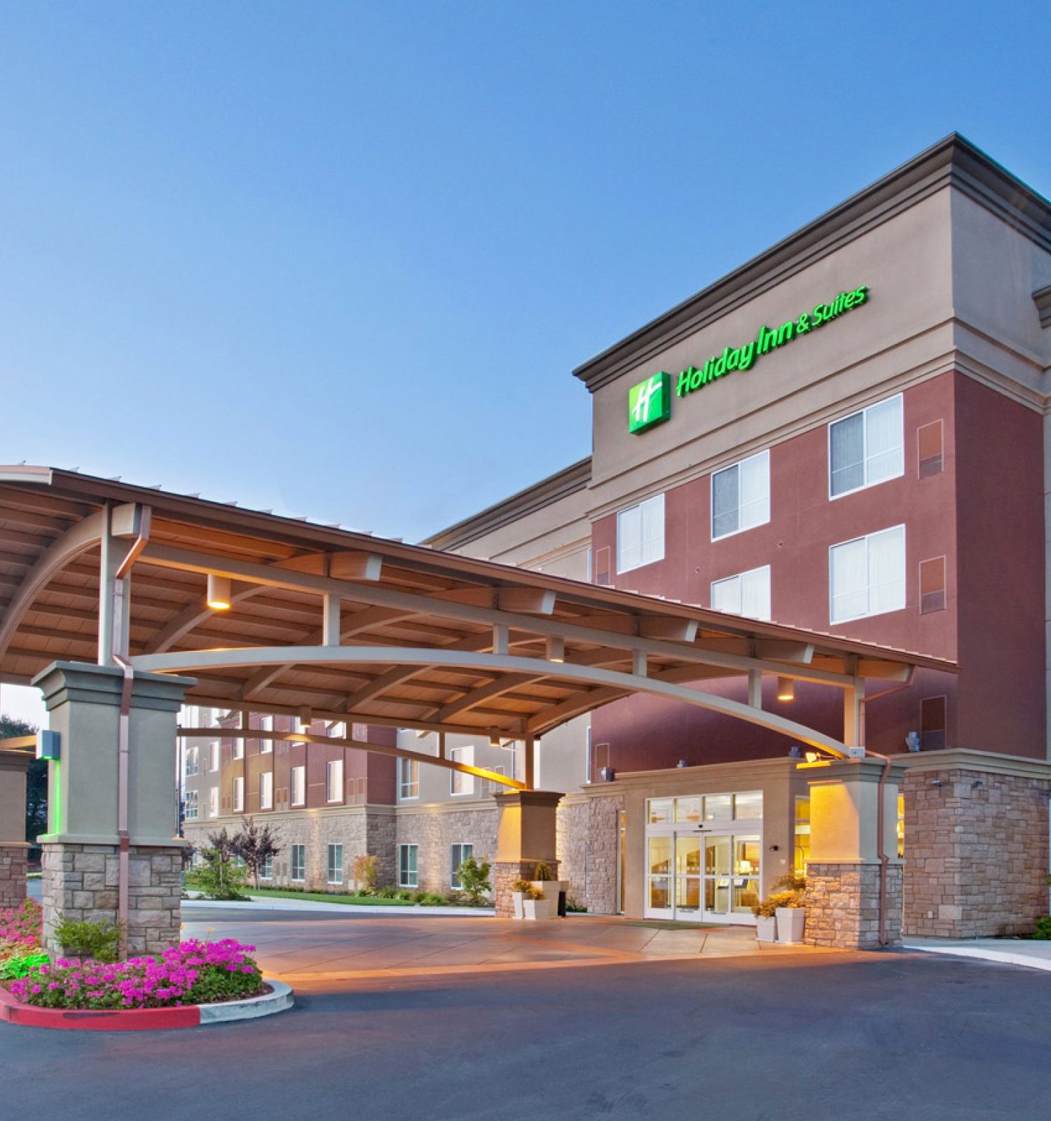 The image shows the exterior of a hotel with a covered entrance and a well-maintained landscape under a clear blue sky at dusk.