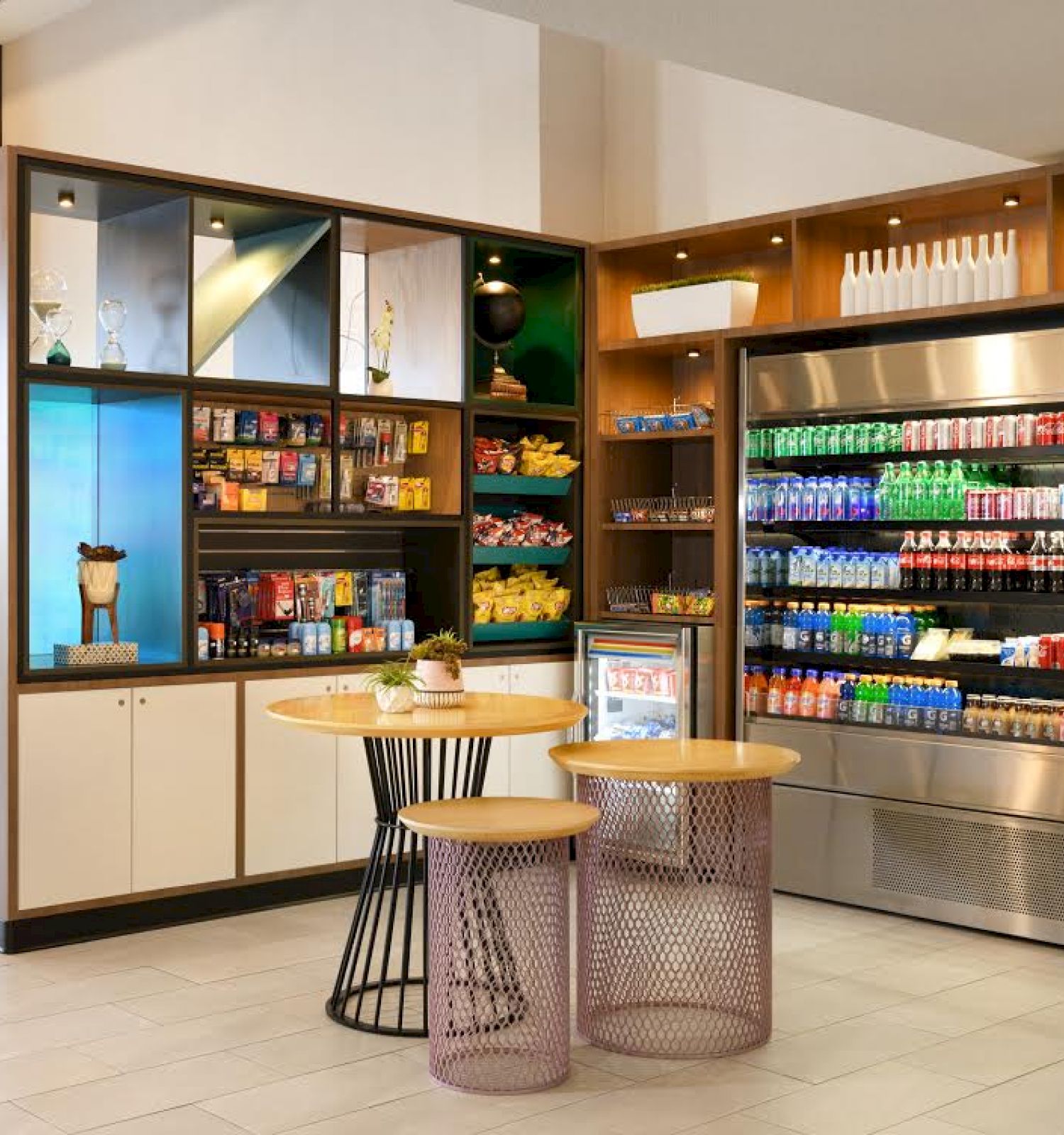 A small convenience snack area with shelves of snacks, a refrigerated section of drinks, and two tables with stools in a well-lit modern space.