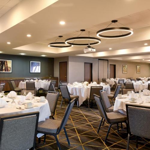 The image shows an elegantly set banquet hall with round tables and chairs, white tablecloths, and modern lighting fixtures hanging from the ceiling.