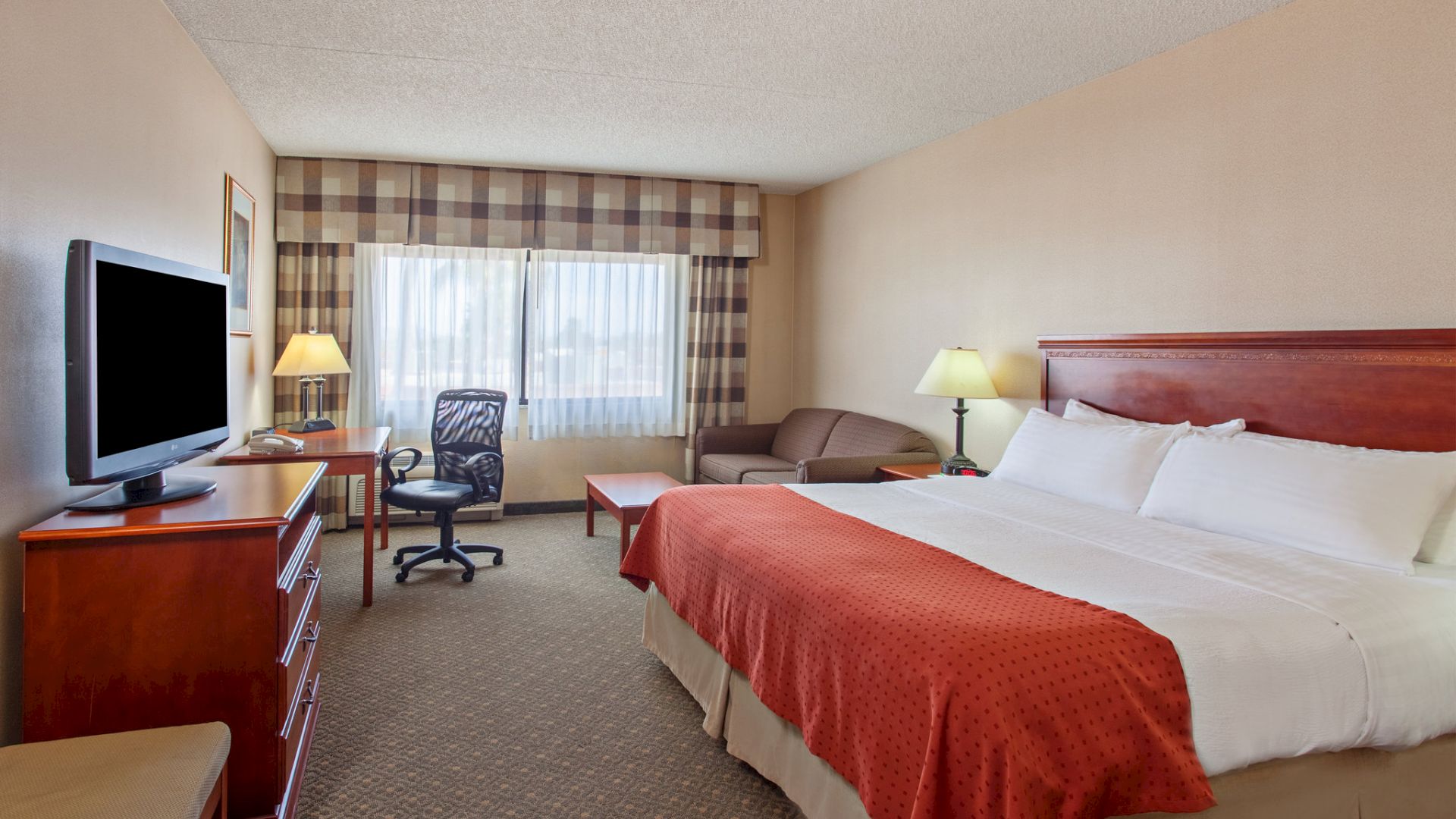 A hotel room with a large bed, television, desk with chair, sofa, and two lamps. The window is covered by curtains, and the room has beige walls.