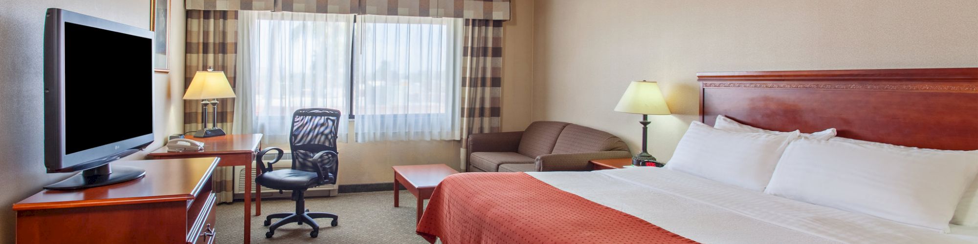 A hotel room with a large bed, television, desk with chair, sofa, and two lamps. The window is covered by curtains, and the room has beige walls.